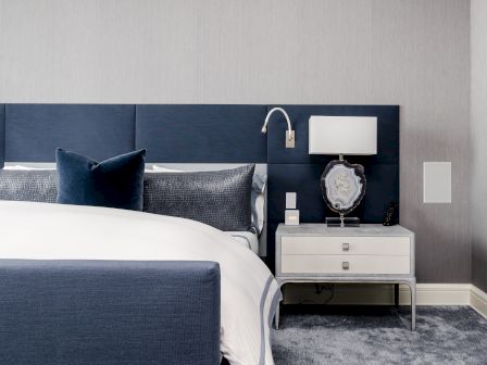 A modern bedroom with a blue headboard, white bedding, and a nightstand featuring a lamp and decorative items, on a gray carpet.