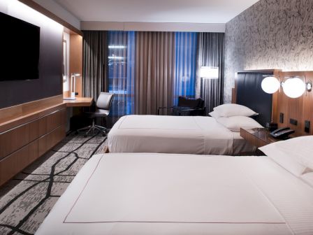 The image shows a modern hotel room with two beds, a flat-screen TV, a desk area, curtains, and stylish decor on the walls.