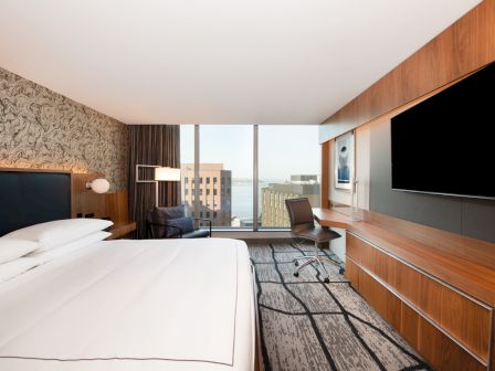 A modern hotel room with a large bed, desk, and flat-screen TV; window views of city buildings and water in the background.