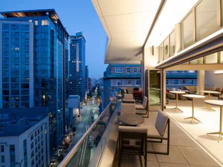 A modern balcony view overlooking a cityscape with tall buildings and a street, featuring seating and tables in an open, well-lit space.