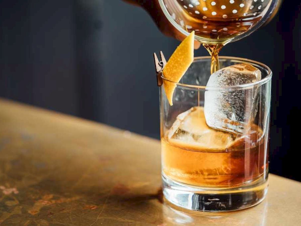 A drink being poured into a glass with ice and a lemon twist garnish on a bar counter.