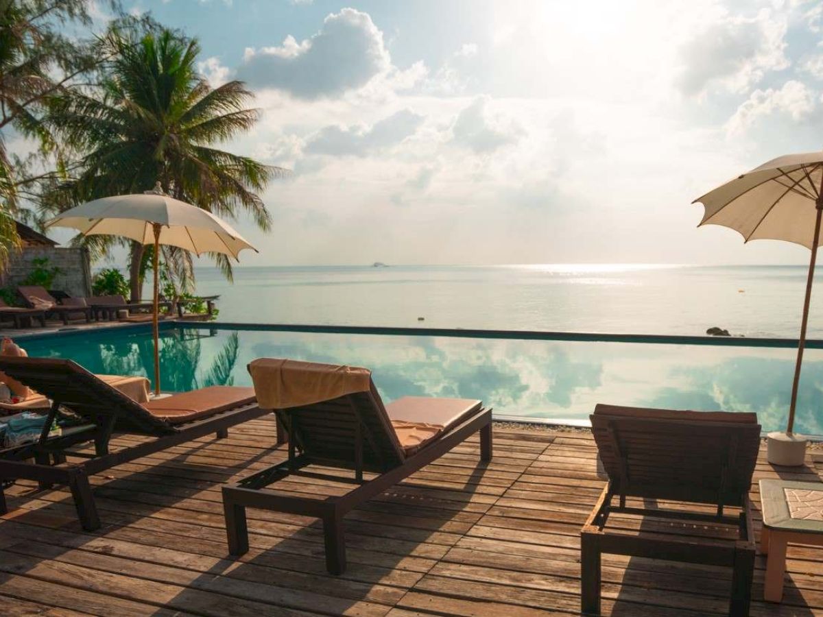 A serene beach scene with lounge chairs, umbrellas, and a palm tree by an infinity pool, overlooking the calm sea and a partly cloudy sky.