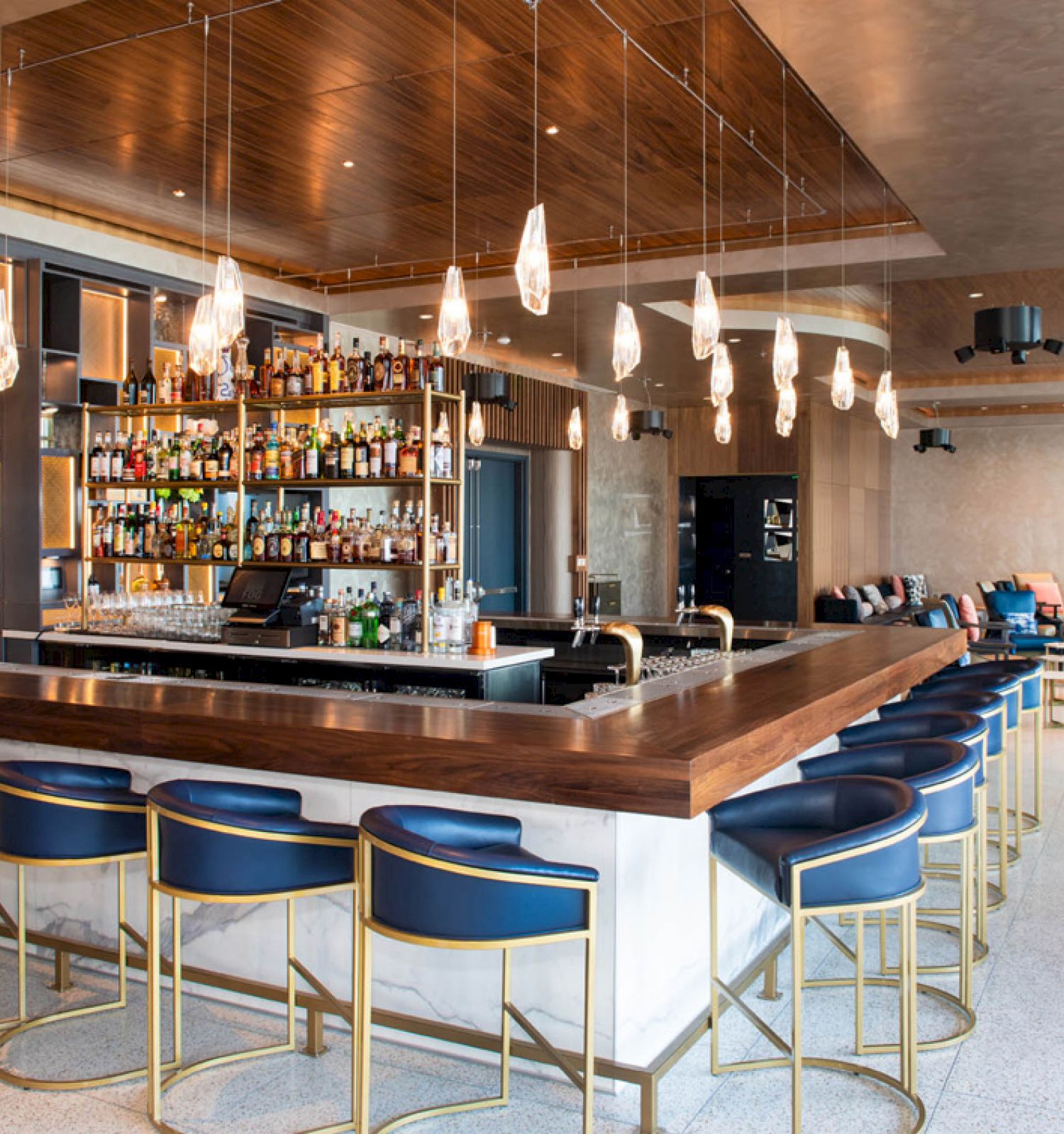 A modern bar with stylish hanging lights, stocked shelves, and blue chairs around a wooden counter in a chic lounge setting.