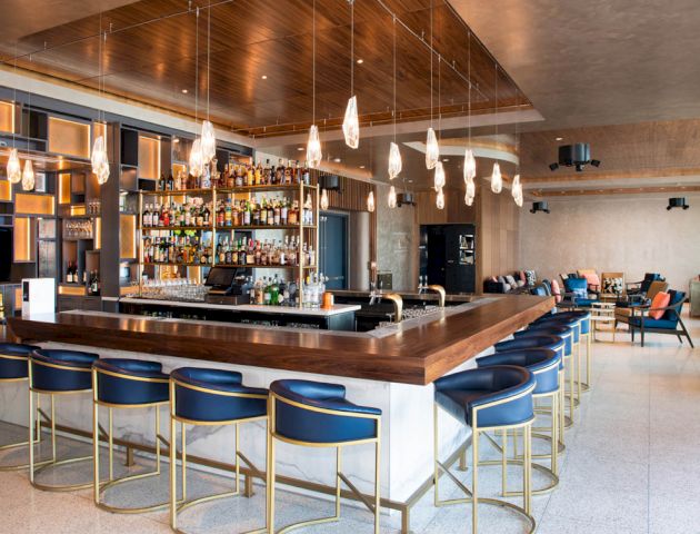 The image shows a modern bar with a wooden counter, blue and gold stools, hanging lights, and shelves stocked with bottles.