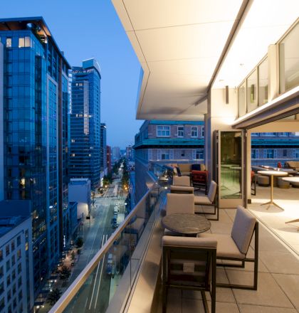 A modern cityscape at twilight with tall buildings, viewed from a sleek balcony with seating and tables, featuring illuminated interiors.