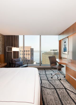 The image shows a modern hotel room with a large bed, a wall-mounted TV, and a desk area by the window overlooking a city view.