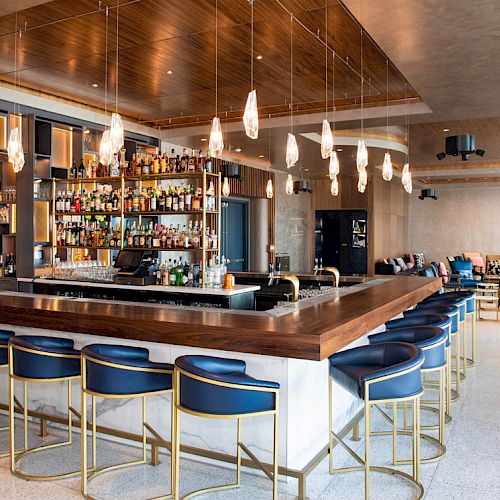 The image shows a modern bar with a wooden counter, shelves of bottles, stylish hanging lights, and blue bar stools in a spacious setting.