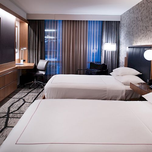 The image shows a modern hotel room with two beds, a wall-mounted TV, curtains, a desk, chair, and stylish lighting fixtures.