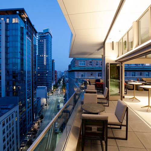 A modern balcony view overlooking a cityscape at dusk, with seating and tables, surrounded by tall, illuminated buildings.