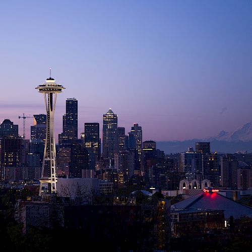 Seattle skyline at dusk with Space Needle and Mount Rainier in the background, under a purple and blue sky.