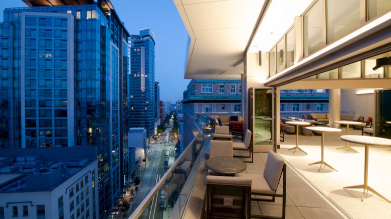 A modern cityscape viewed from a stylish balcony with tables and chairs, overlooking tall buildings and a street below at dusk.