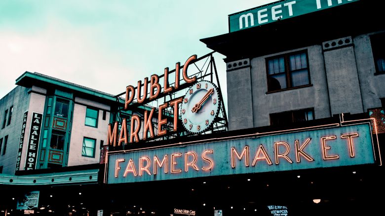 The image shows a neon-lit sign reading "Public Market" and "Farmers Market" on a building, with a clock and surrounding structures.