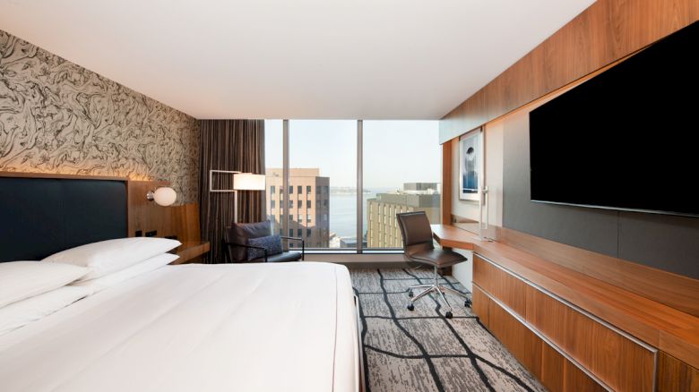 A modern hotel room with a large bed, desk, chair, and flat-screen TV, overlooking city buildings and water through large windows.