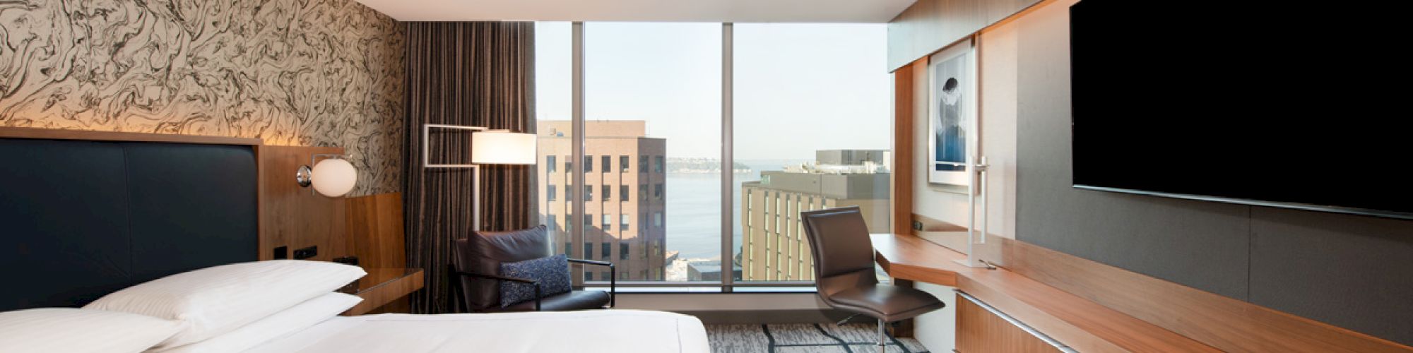 A modern hotel room with a large bed, a wall-mounted TV, a desk, and a window offering a view of a cityscape.
