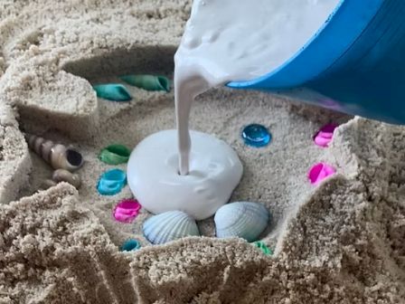 A creamy substance is being poured into a sand mold surrounded by colorful shells and stones, creating a decorative beach craft.
