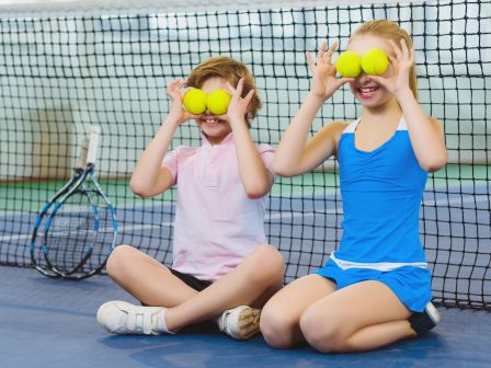 Two children sitting on a tennis court hold tennis balls over their eyes, smiling. A net and rackets are in the background.