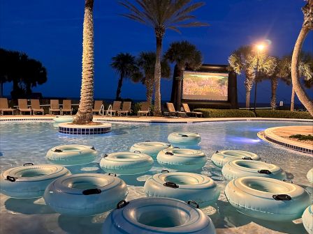 A pool scene at night with inflatable tubes, palm trees, and an outdoor movie screen set up in the background.