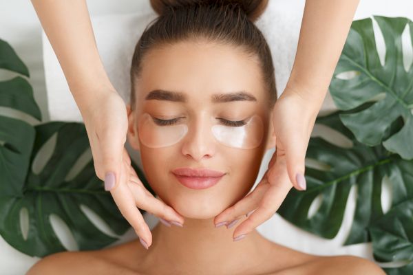A person is receiving a facial treatment with under-eye patches applied, surrounded by green leaves, while hands gently touch their face.