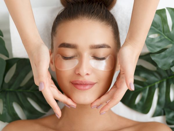 A person is receiving a facial treatment with under-eye patches applied, surrounded by green leaves, while hands gently touch their face.