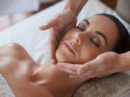 A peaceful woman receives a facial massage; hands cradle her head and neck as she relaxes with closed eyes, enjoying gentle spa treatment.