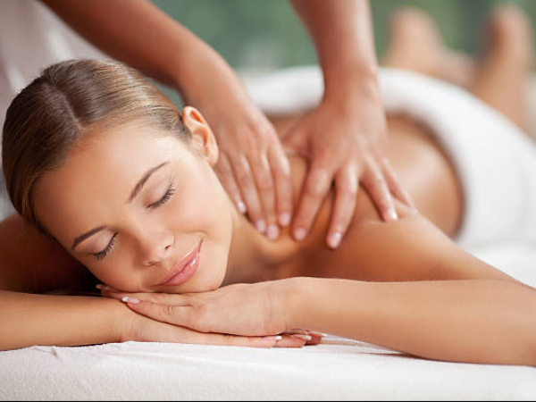 A woman enjoys a relaxing back massage while lying face-down with eyes closed and hands resting, as a therapist kneads her shoulders.