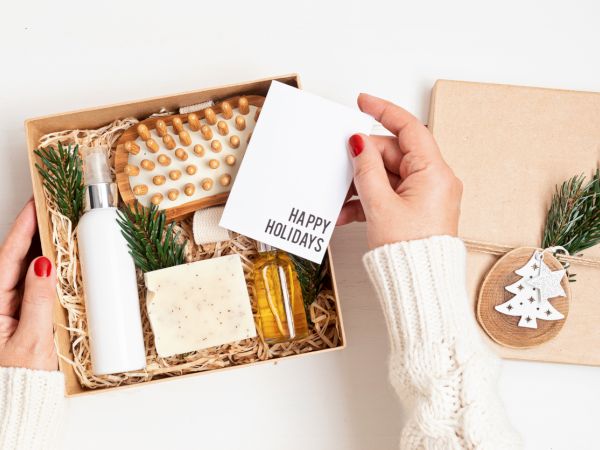 A holiday gift box opens with a bottle, soap, snacks, and a &ldquo;Happy Holidays&rdquo; card; festive sprigs and a small Christmas ornament on the lid.