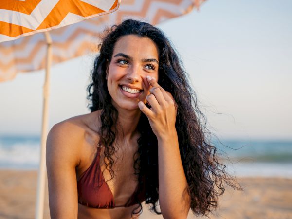 A person smiling on the beach under an umbrella while applying sunscreen.