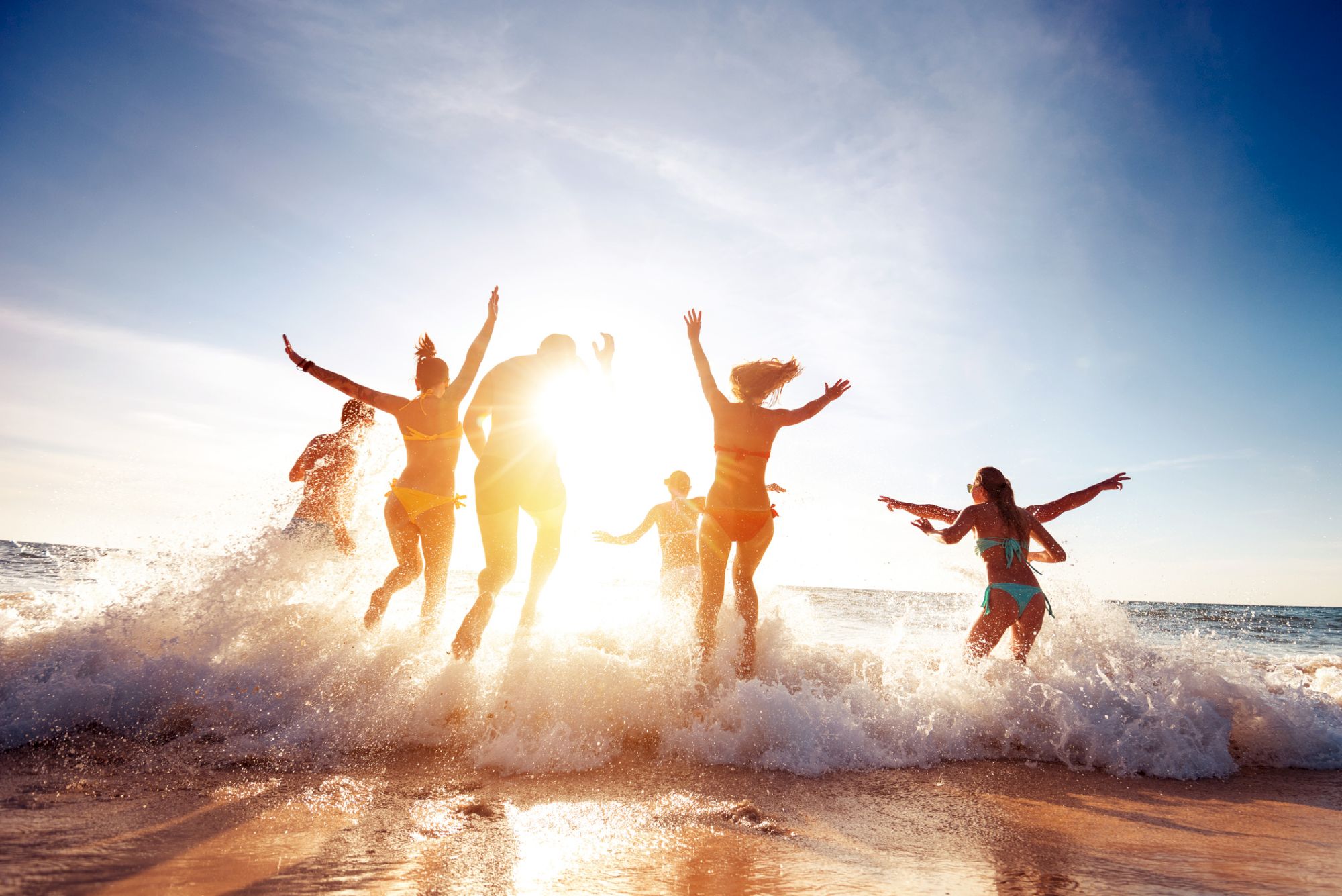 A group of people joyfully playing in the ocean waves with the sun shining brightly behind them, creating a lively summer beach scene.