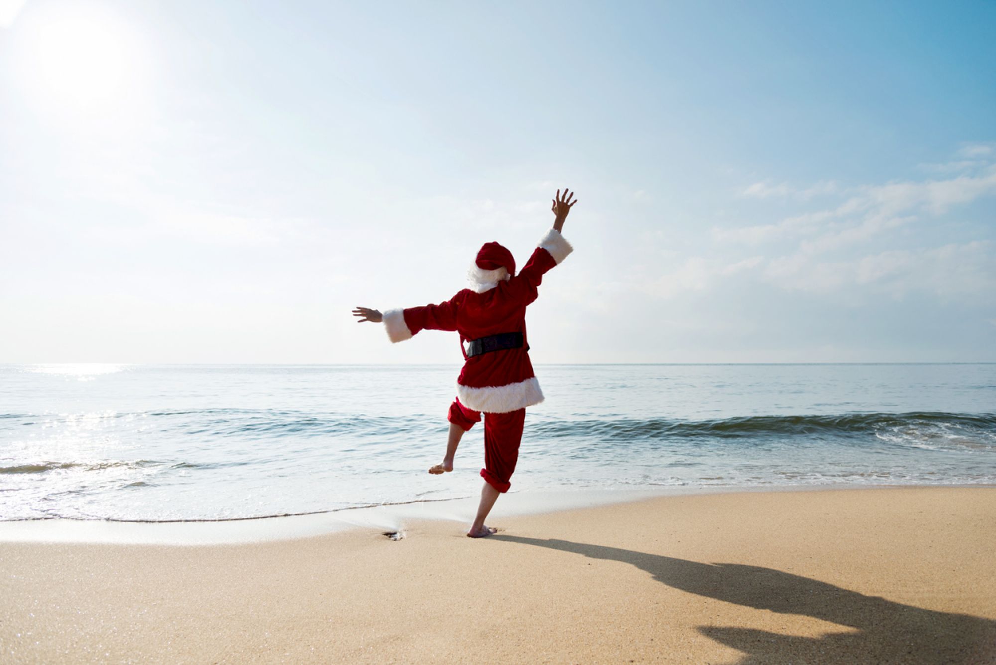 A person in a red outfit joyfully dances on a sunny beach near the sea, throwing arms up as they balance on one leg with a long shadow.