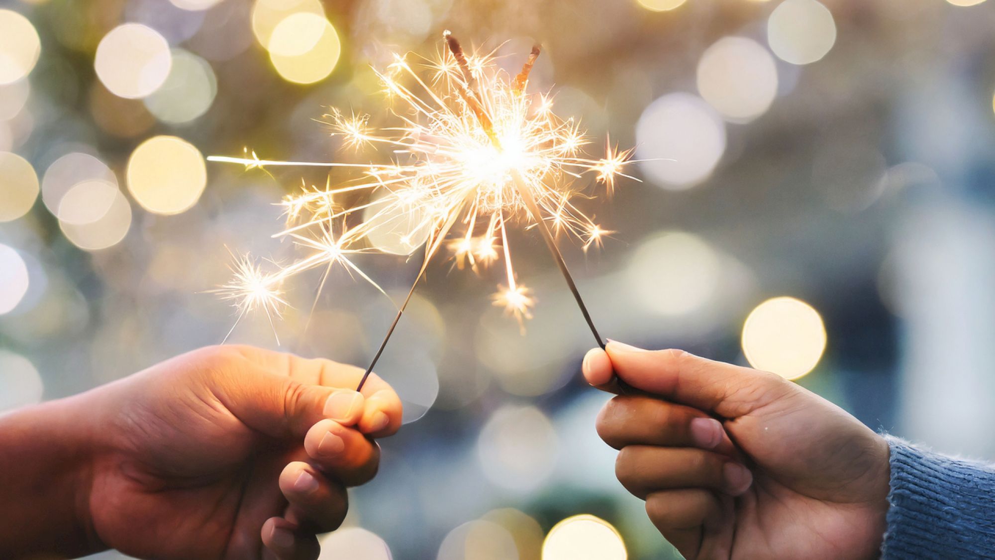 Two people spark a celebration by lighting sparklers, clinking them together as bright bokeh lights glow in the background.