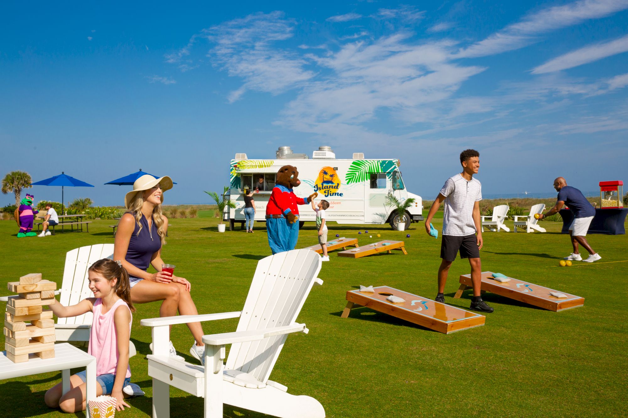 People enjoying outdoor games on a grassy field with cornhole, Jenga, and a food truck in the background under a clear blue sky.