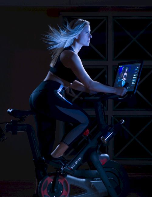 A person exercises on a Peloton stationary bike, focusing on a display screen in a dimly lit room.
