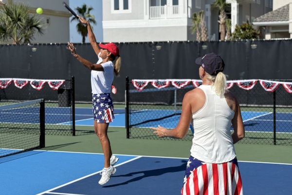 Two women playing tennis on an outdoor court, both wearing patriotic-themed outfits and hats.