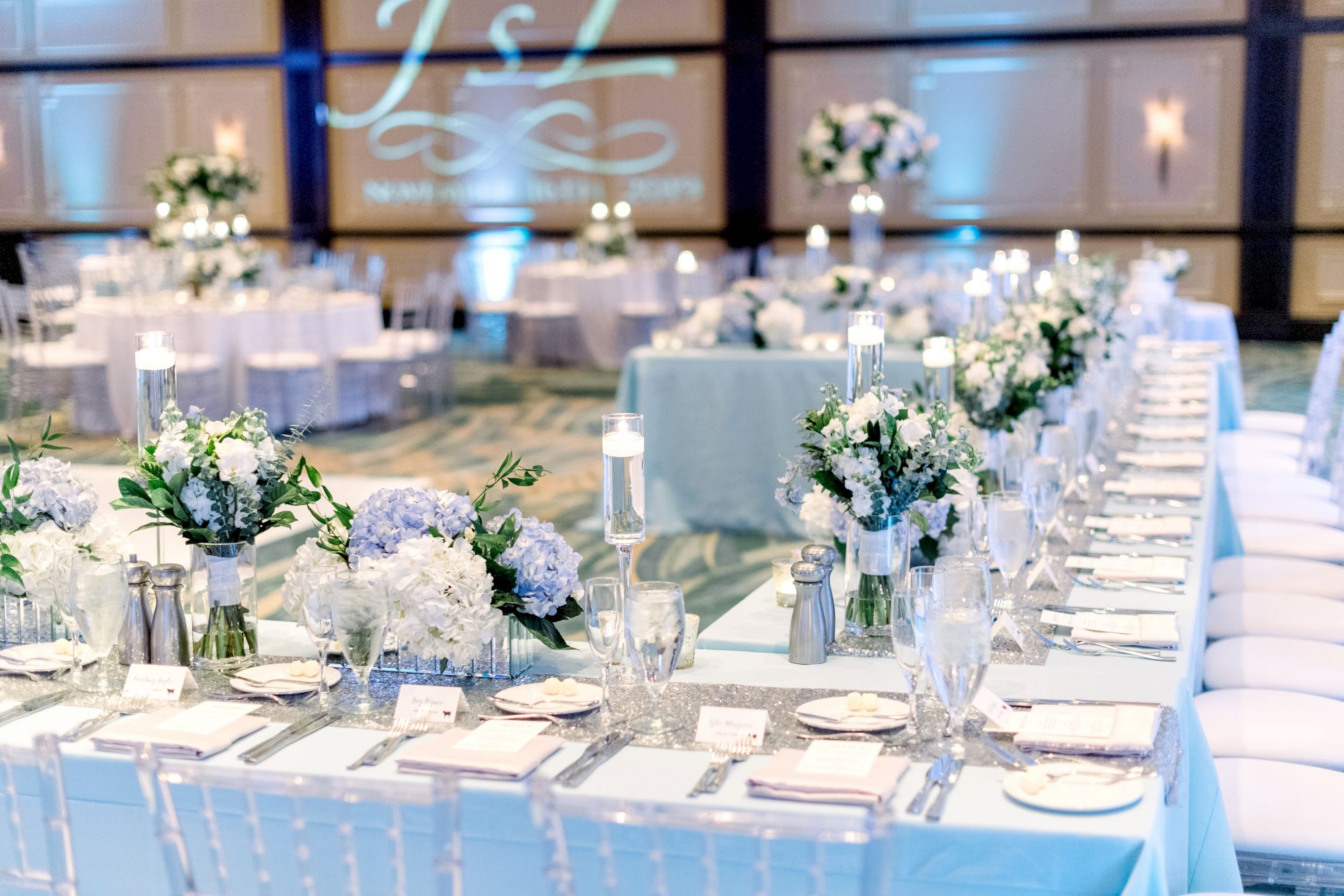 Elegant wedding reception with a long decorated table, floral centerpieces, candles, and white chairs in a softly lit venue.