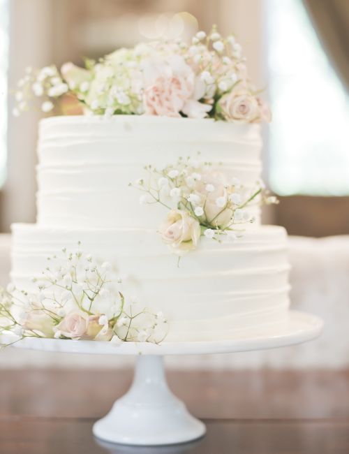 A two-tiered white cake adorned with delicate pink and white flowers on a white cake stand, set against a soft, blurred background.
