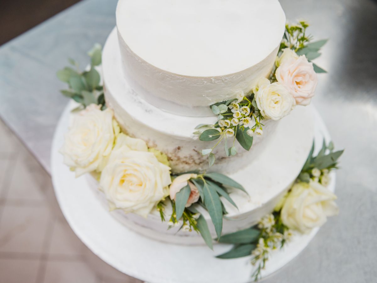 A white three-tier cake decorated with white roses and green leaves is displayed on a stand in this image.