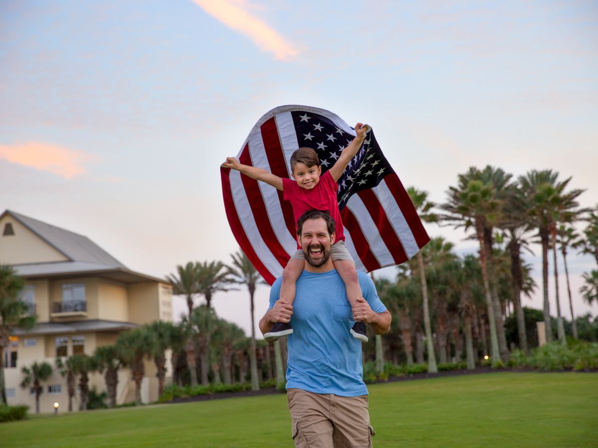 A man carries a child on his shoulders, holding an American flag, with palm trees and a building in the background.