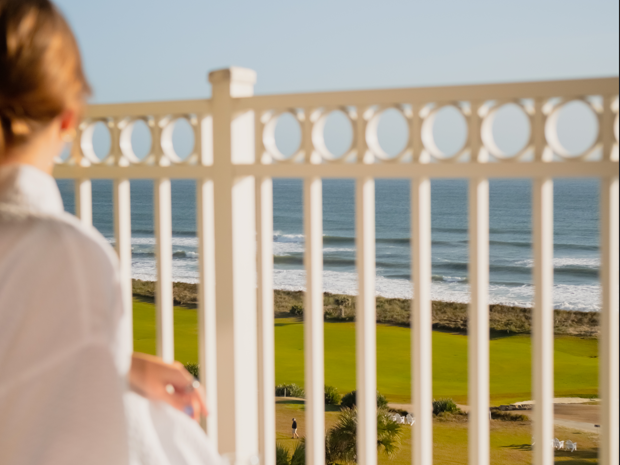 A woman in white sits on a seaside balcony, looking out at the ocean beyond a garden and colonnade.