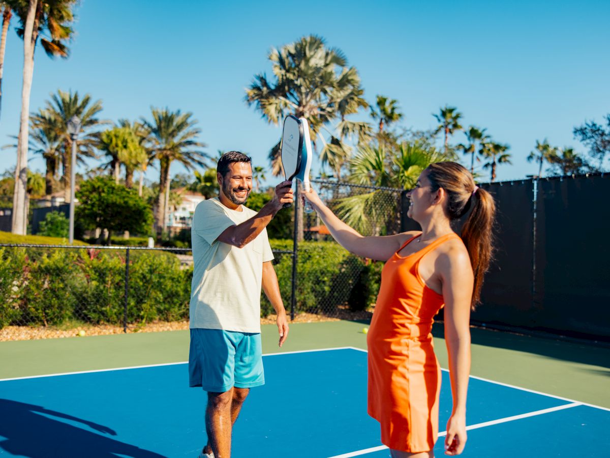 Two people on a tennis court smiling and touching tennis racquets, surrounded by palm trees and a clear blue sky.