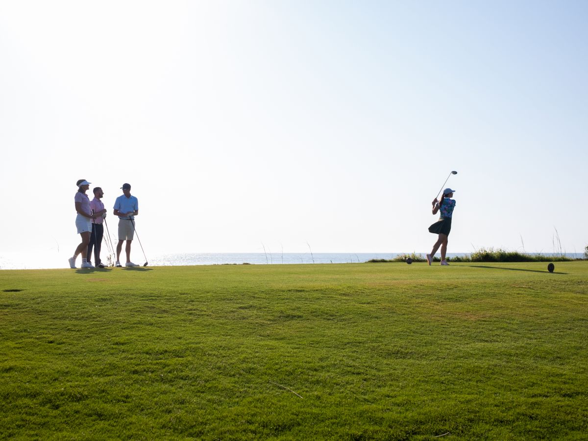 A group of golfers plays on a sunny seaside course, with one swinging a club while others stand and watch nearby.