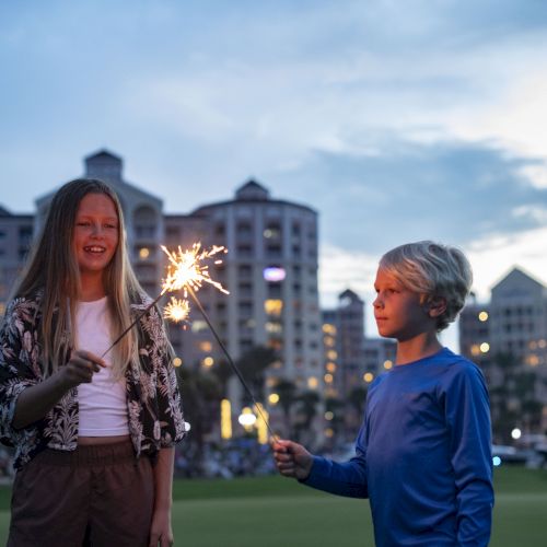 Two kids smile while holding sparklers in a city park, with tall buildings glowing in the evening sky behind them.